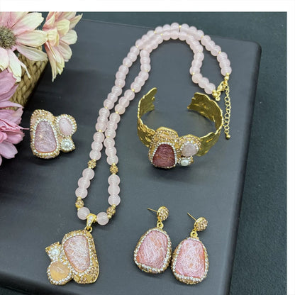 A jewelry set displayed on a dark gray surface, featuring a gold-tone cuff bracelet, matching pendant necklace, and two matching drop earrings. The jewelry is decorated with light pink beads, gold-tone filigree, and pink stone inlays.