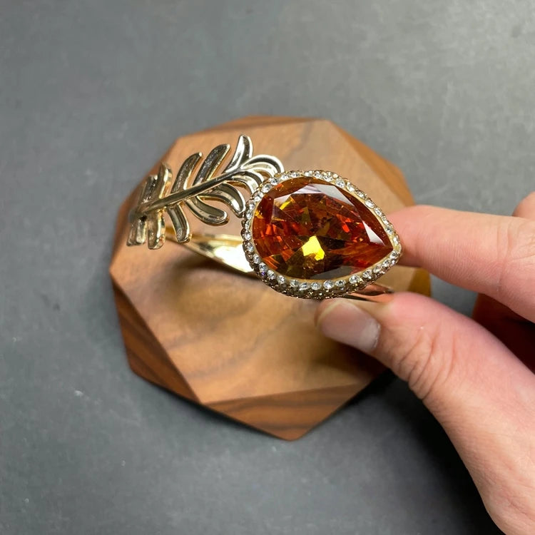 A gold-toned leaf-shaped cuff bracelet with a large teardrop-shaped amber gemstone and a halo of small clear stones, held by a hand over a faceted wooden block on a gray surface.