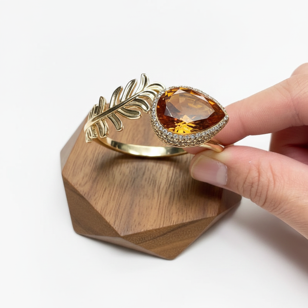 A gold leaf-shaped cuff ring with a large teardrop-shaped amber gemstone and a halo of small clear stones, held by a hand over a faceted wooden display block.