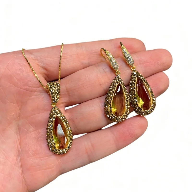 Gold-tone jewelry set including a teardrop pendant necklace and matching drop earrings, displayed on a person's hand against a white background.