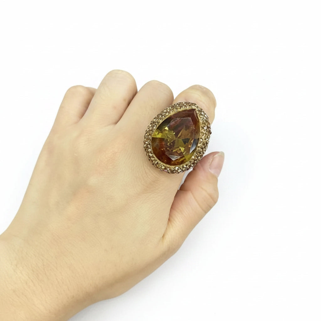 Hand wearing a large ring with a teardrop-shaped gemstone on a light gray background